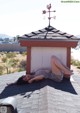 A woman laying on top of a roof next to a weather vane.